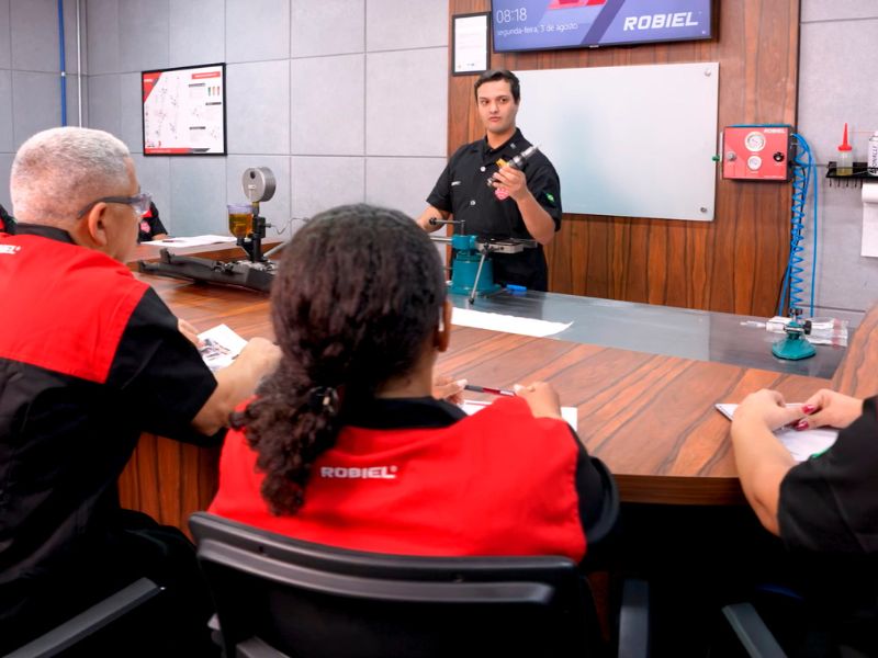 Sala de treinamento presencial da ROBIEL com instrutor demonstrando componente diesel para alunos sentados em bancada de madeira, ambiente técnico iluminado, fotografia realista